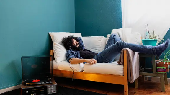 Ein Mann liegt auf einem Sofa, daneben ein Plattenspieler, und hört Musik über Kopfhörer.