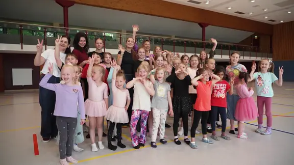 In einer Turnhalle stehen Kinder, Jugendliche und Erwachsene einer Tanzgruppe und winken in die Kamera.