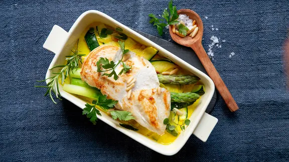 Eine Auflaufform mit Spargel-Zucchini-Auflauf mit Hähnchenfilet stehen auf einem Tisch.