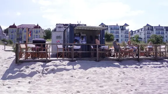 Ein kleiner Container, eine Bar steht am Strand, davor Gäste.