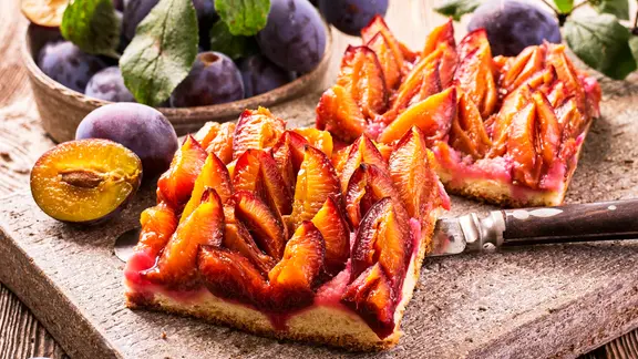 Zwei Stücke Zwetschgenkuchen liegen auf einem Holzbrett, dahinter und daneben weitere Zwetschgen.
