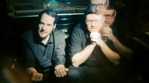 Gisbert zu Knyphausen und Kai Schumacher sitzen nebeneinander vor einem geöffneten Klavier. Das Bild ist teilweise mehrfach belichtet, wodurch das Gesicht Schumachers mehrfach sichtbar wird.
