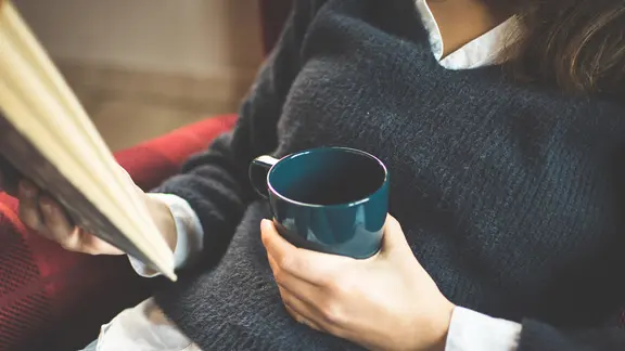 Eine junge Frau im blauen Pullover sitzt gemütlich im Sessel und hält einen Kaffeebecher und ein Buch in ihren Händen.