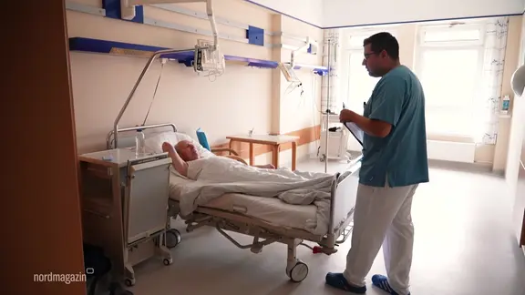 Ein Pfleger steht vor dem Bett eines Patienten auf einer Station in einem Krankenhaus.