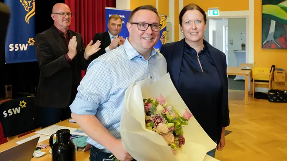 Ein Mann steht neben einer Frau in einem Raum mit anderen Menschen und hält einen Blumenstraus in den Händen.