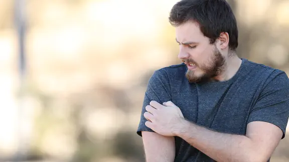 Ein Mann im dunkelblauen T-Shirt in der Natur kratzt sich am Arm und schaut gereizt