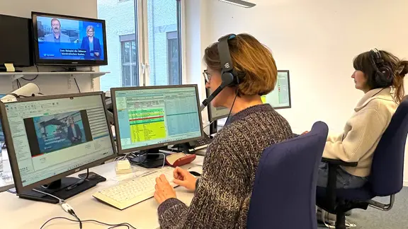 Zwei Frauen mit Headsets sitzen vor Monitoren mit dem Fernsehbild.