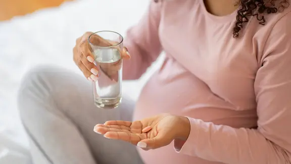 Eine Schwangere Frau hält ein Wasserglas in der einen und eine Tablette in der anderen Hand.