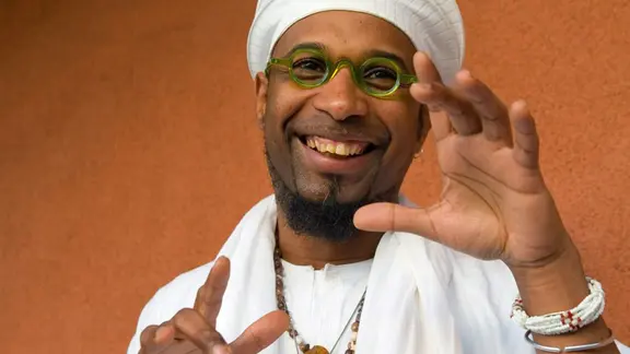 Omar Sosa lächelt und steht in weißer Kleidung und mit einer grünen Brille auf vor einer orangefarbenen Wand. Er hält die Hände in einer offenen, gestikulierenden Pose und trägt mehrere Halsketten und Armbänder.