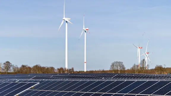 Im Vordergrund ist eine Photovoltaik-Anlage zu sehen, im Hintergrund stehen Windräder.