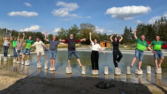Eine Gruppe von zehn Personen steht auf Holzpfosten am Rand eines Naturbad-Beckens. Sie balancieren, heben jeweils einen Arm und ein Bein, lächeln und wirken fröhlich.