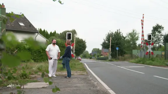 Zwei Personen unterhalten sich am Straßenrand vor einem Bahnübergang.