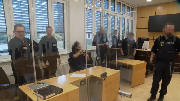 Ein Angeklagter sitzt um Gericht und Sicherheitspersonal steht hinter ihm.