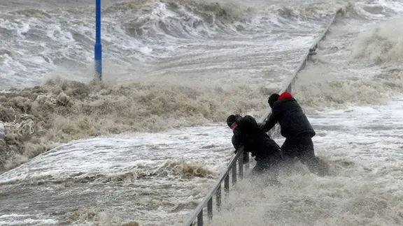 Zwei Männer halten sich an einem Geländer an der vom Sturm überspülten Uferpromenade in Dagebüll fest