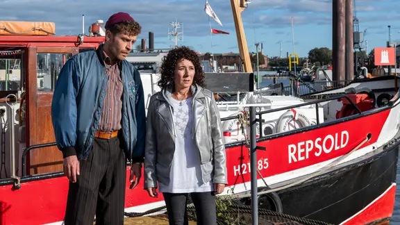 Eine Frau und ein Mann stehen vor einem Schiff, das im Hafen liegt.