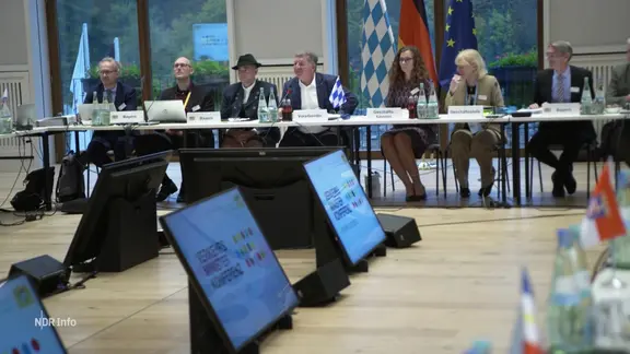 Menschen an Konferenztischen, im Hintergrund Länderfahnen.