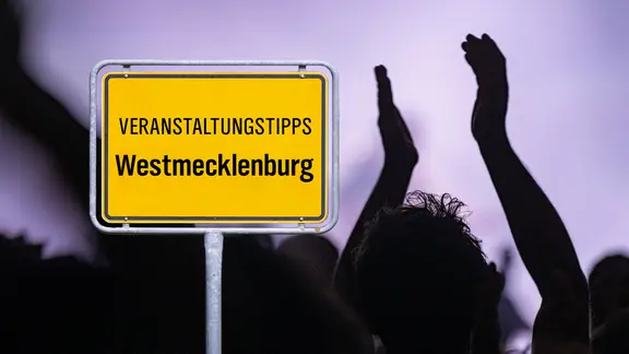 Ein Schild mit der Aufschrift "Veranstaltungstipps in Westmecklenburg" vor Personen.