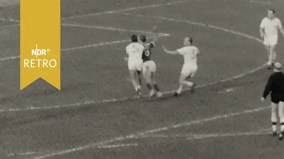 Szene aus einem Feldhandballspiel 1963: Ein Angreifer des Büdelsdorfer TSV beim Torwurf gegen zwei Verteidiger des VfL Wolfsburg
