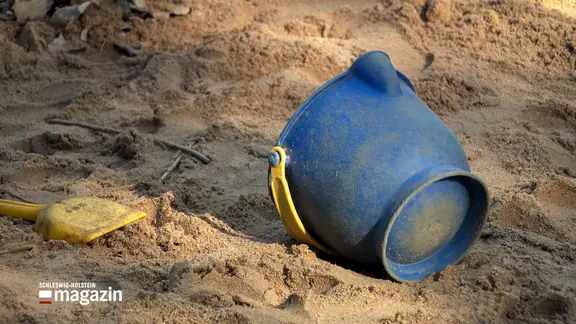 Ein kleiner blauer Plastikeimer und eine gelbe Schaufel liegen in einer Sandkiste in einer Kita.