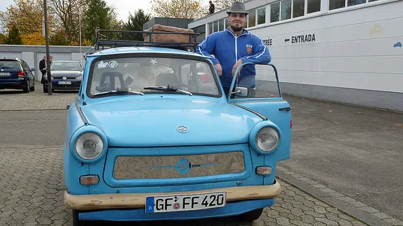 Ein Mann steht in DDR-Trainingsjacke und Hut an einem hellbaluen Trabbi.