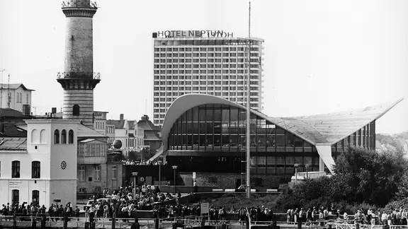 Blick auf den alten Leuchtturm und den Teepott an der Seepromenade des Ostseebades Warnemünde in Rostock, aufgenommen im Juli 1972. Dahinter ist das 1971 in Betrieb genommene Hotel "Neptun" zu sehen. Der 36,90 Meter hohe Leuchtturm wurde 1898 in Betrieb genommen, der Teepott wurde 1967 erbaut.