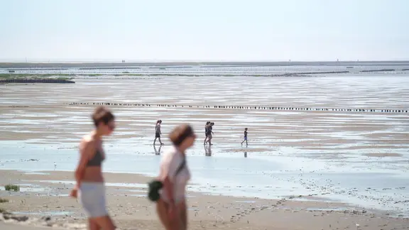 Zwei Frauen im Vordergrund gehen am Strand entlang, im Hintergrund laufen vier Personen bei Ebbe durchs Wasser.