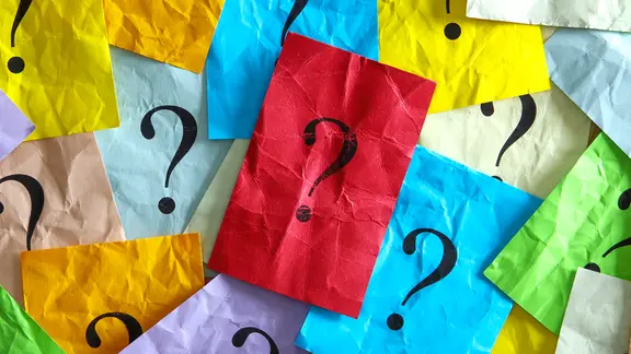 Mehrere bunte, zerknitterte Papierblätter liegen übereinander, jedes mit einem schwarzen Fragezeichen in der Mitte. Ein rotes Blatt mit Fragezeichen ist zentral und sticht hervor.