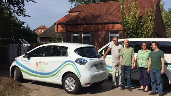 Zu sehen sind zwei Elektro-Autos und vier Personen die vor einem der Autos stehen und Richtung Kamera gucken.