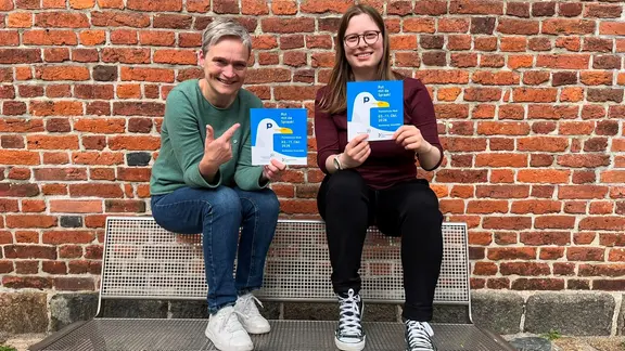 Zwei Frauen sitzen auf einer Bank und halten ein Plakat in den Händen.