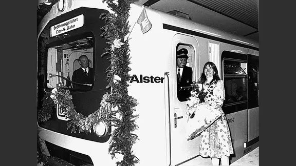 Ein blumengeschmückter S-Bahn-Zug mit Hamburg-Fahne. Helga Feddersen steht auf dem Bahnsteig, der Lokführer schaut aus der Tür.