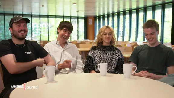 Drei Männer und eine Frau sitzen in einer Cafeteria zusammen am Tisch und lächeln in die Kamera.