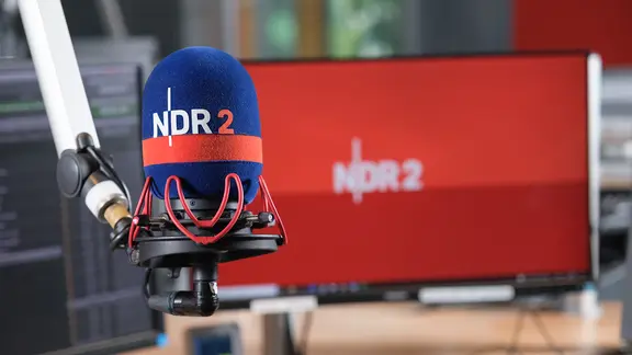 Blick auf ein Mikrofon mit NDR 2 Schützer im NDR 2 Studio mit einem Bildschirm im Hintergrund, auf dem auf rotem Grund ein weißes NDR 2 Logo zu sehen ist.