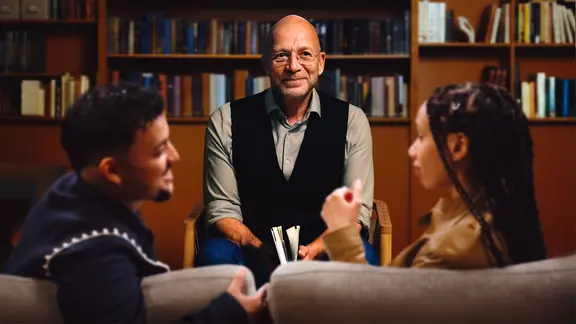 Therapeut Eric Hegmann sitzt vor einem Bücherregal und spricht mit einem Paar auf dem Sofa