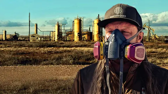 Ein Mann mit Atemmaske aus Gummi mit Filterkammern links und rechts vor einer flachen Landschaft mit Gas-Silos in den USA.