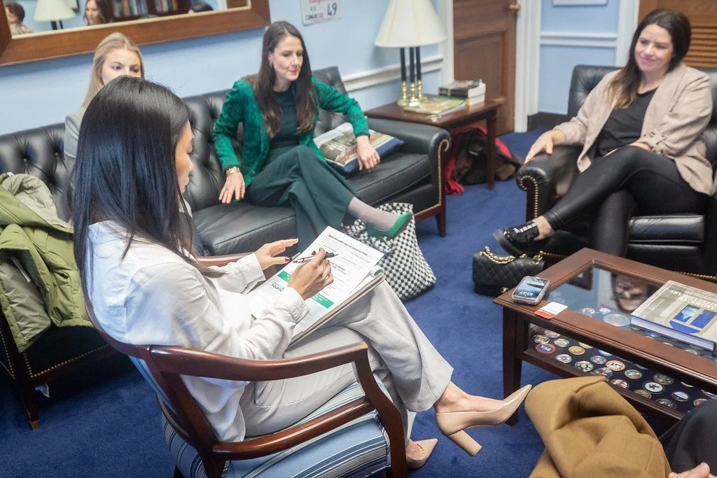 Four women sitting in a room engaged in a discussion; one woman is writing on a clipboard while the others listen.