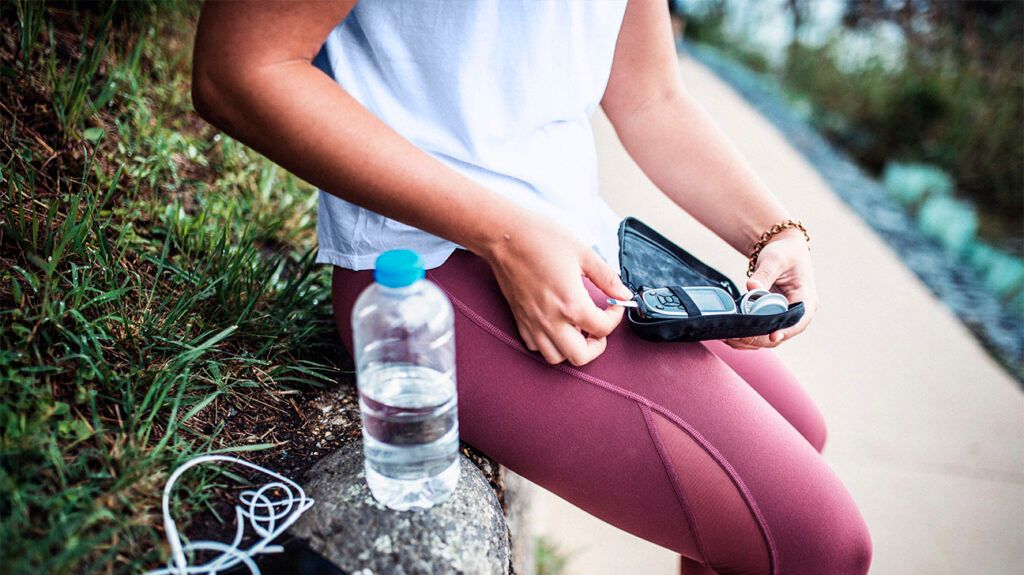 Close-up of a person in sports clothing sitting on a wall and pulling a blood glucose monitor from its case.