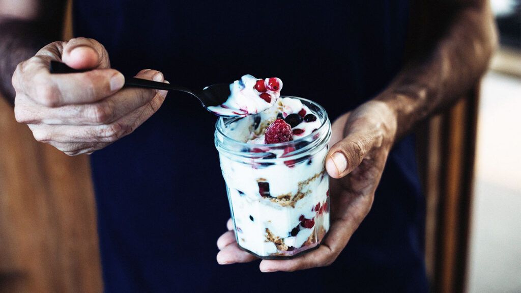 A man holding a jar of glass jar containing yoghurt, berries, and granola. Eating it with a spoon.