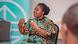 Nouratou Zato, wearing a brightly colored dress, sits on a chair and gesticulates with her hands.