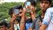 Several young men stand outdoors holding smartphones, taking photos or videos. The focus is on their hands and devices, with a blurred green background
