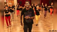A young man leading a dance class in a dance hall. Behind him is a group of women, all their faces are blurred.