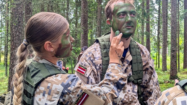 Während eines freiwilligen militärischen Trainings für Schülerinnen und Schüler in Lettland trägt eine Teilnehmerin dem quer-Reporter Philipp Potthast Tarnschminke auf.  | Bild: BR/Viola Nowak