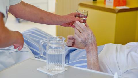 Im Vordergrund übergibt eine Hand ein Glas Wasser in die Hand eines alten Menschen, der in einem Krankenhausbett liegt. Auf dem Beistelltisch des Krankenbettes steht eine Karaffe.