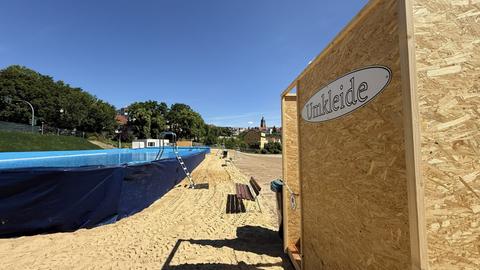 Ein rechteckiges, oberirdisches Schwimmbecken steht auf einer mit Sand bedeckten Fläche, daneben führt eine Metallleiter ins Wasser. Rechts im Vordergrund befindet sich eine hölzerne Kabine mit dem Schild „Umkleide“, im Hintergrund sind Bäume und Gebäude unter blauem Himmel zu sehen.