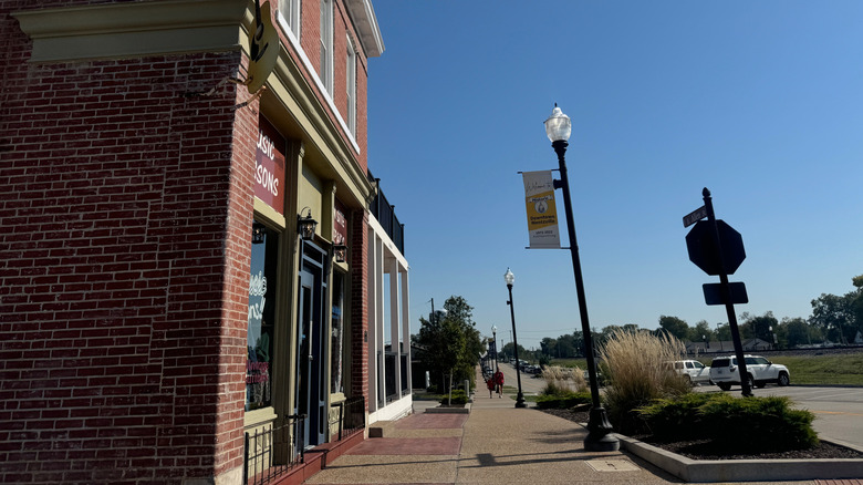 People walking on the sidewalk beside a still street lined with cars, trees, and a red-brick building on a clear day in Wentzville, MO