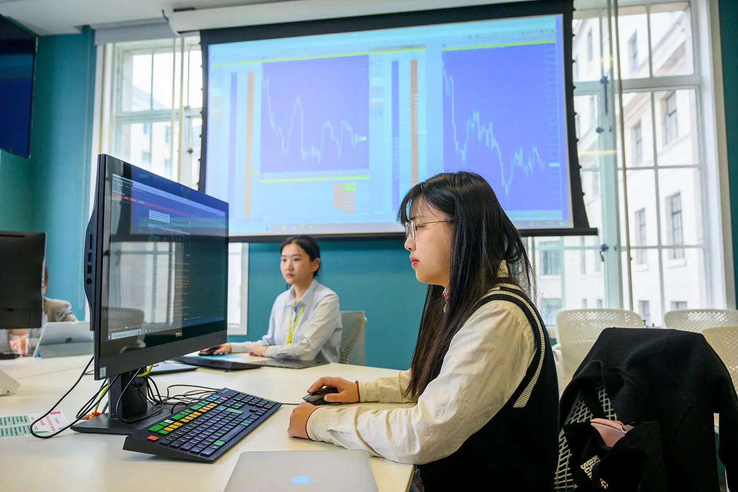 Two people focus on computer monitors displaying financial graphs in an office. The atmosphere is analytical and professional.