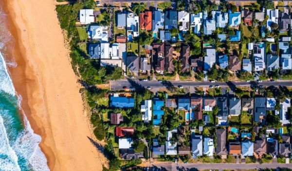 Aerial view of a beachside suburb with roofs, gardens and the sand visible.