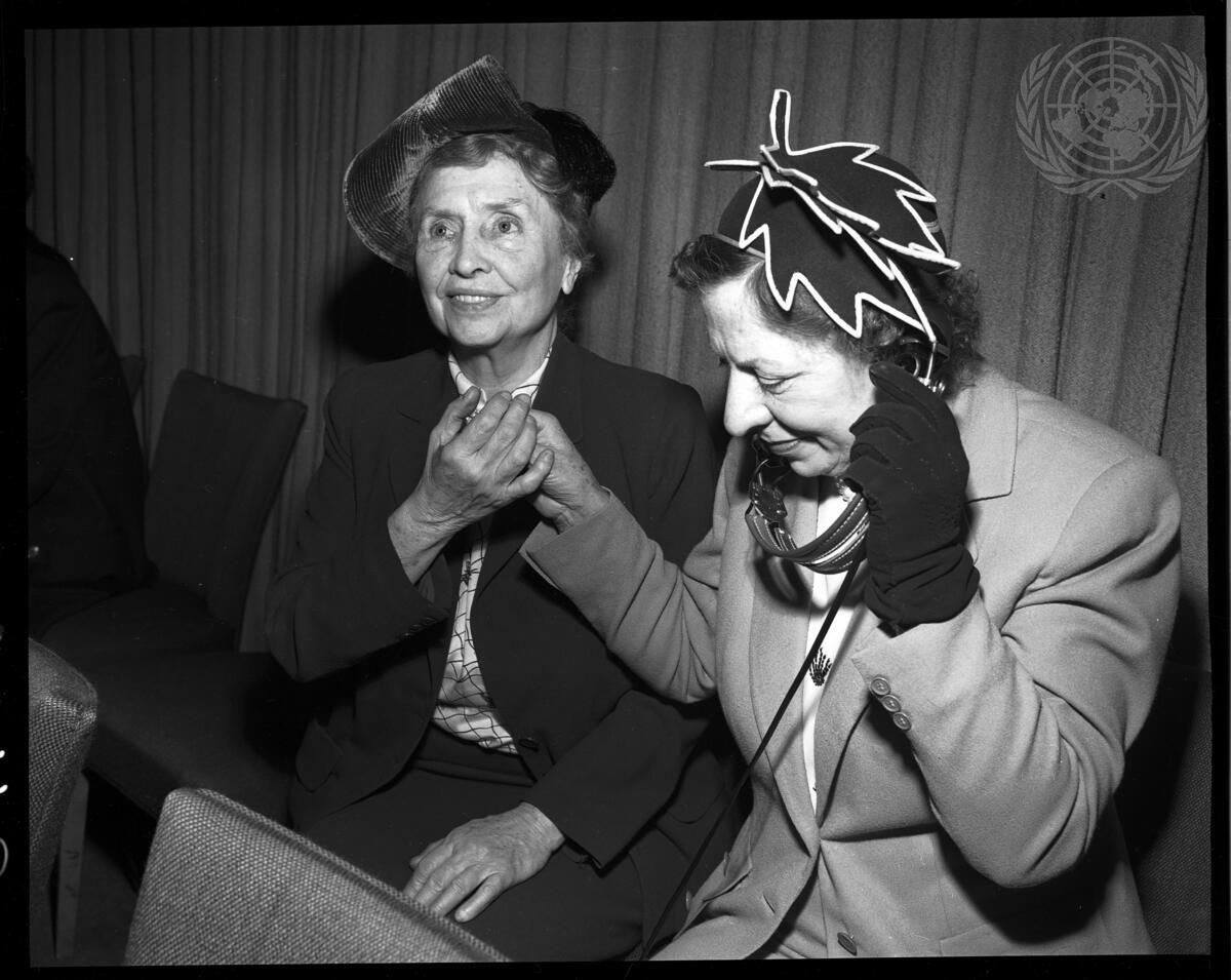 Two women sit together; one wears headphones, while the other holds her hand.
