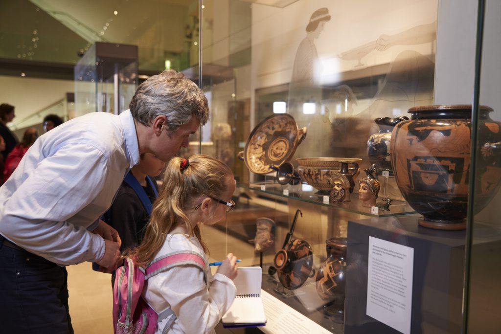 An adult and a child observe ancient pottery in a museum display, taking notes.