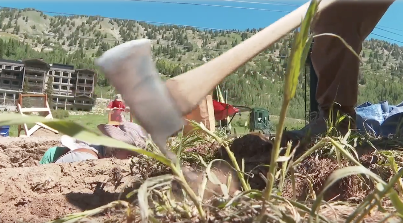 Close-up of an axe chopping soil near a mining town, with mountains and people in the background.