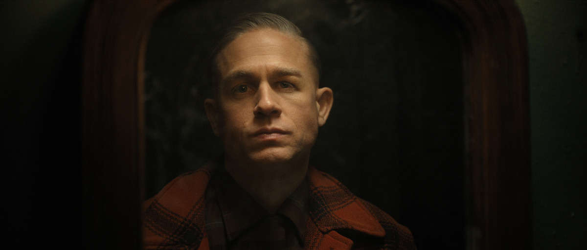 A man with a serious demeanor and visible facial scars sits in a dimly lit area, his presence filling the environment with intensity.